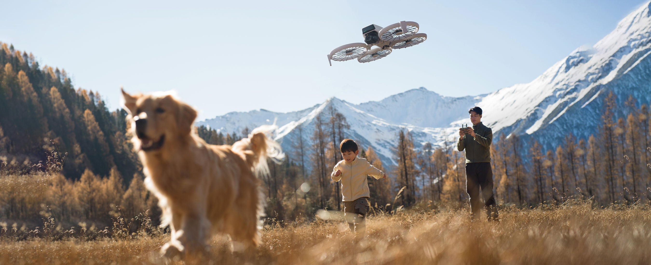 A golden retriever runs with a child in a golden, grassy field with a snowy mountain backdrop while a person records them using the DJI RC 2 and DJI Flip.