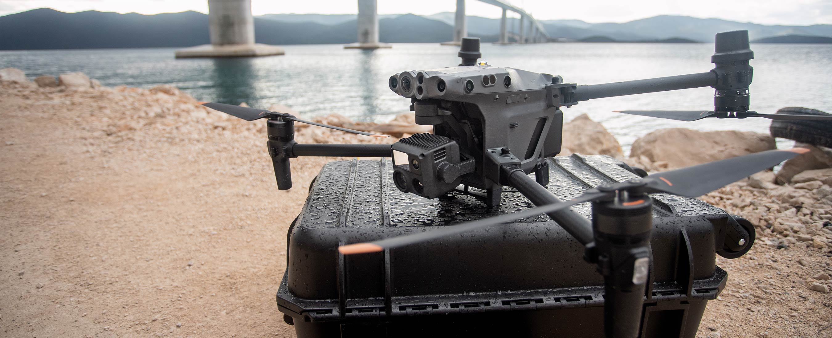 A Matrice 30T displayed on a hardshell case which is resting upon the ground with water, mountains, and a bridge in the background.