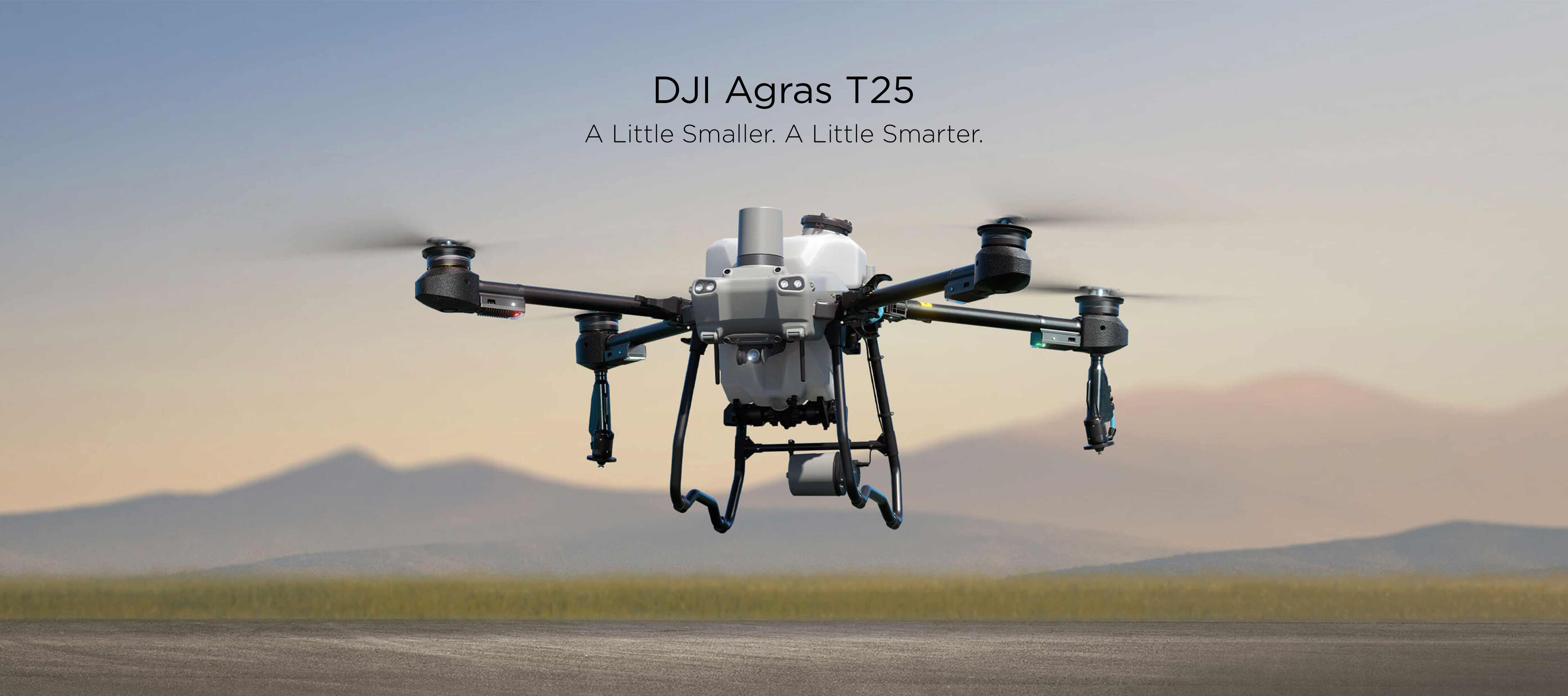 A DJI Agras T25 hovers over sandy terrain with out-of-focus grass and mountains in the background.