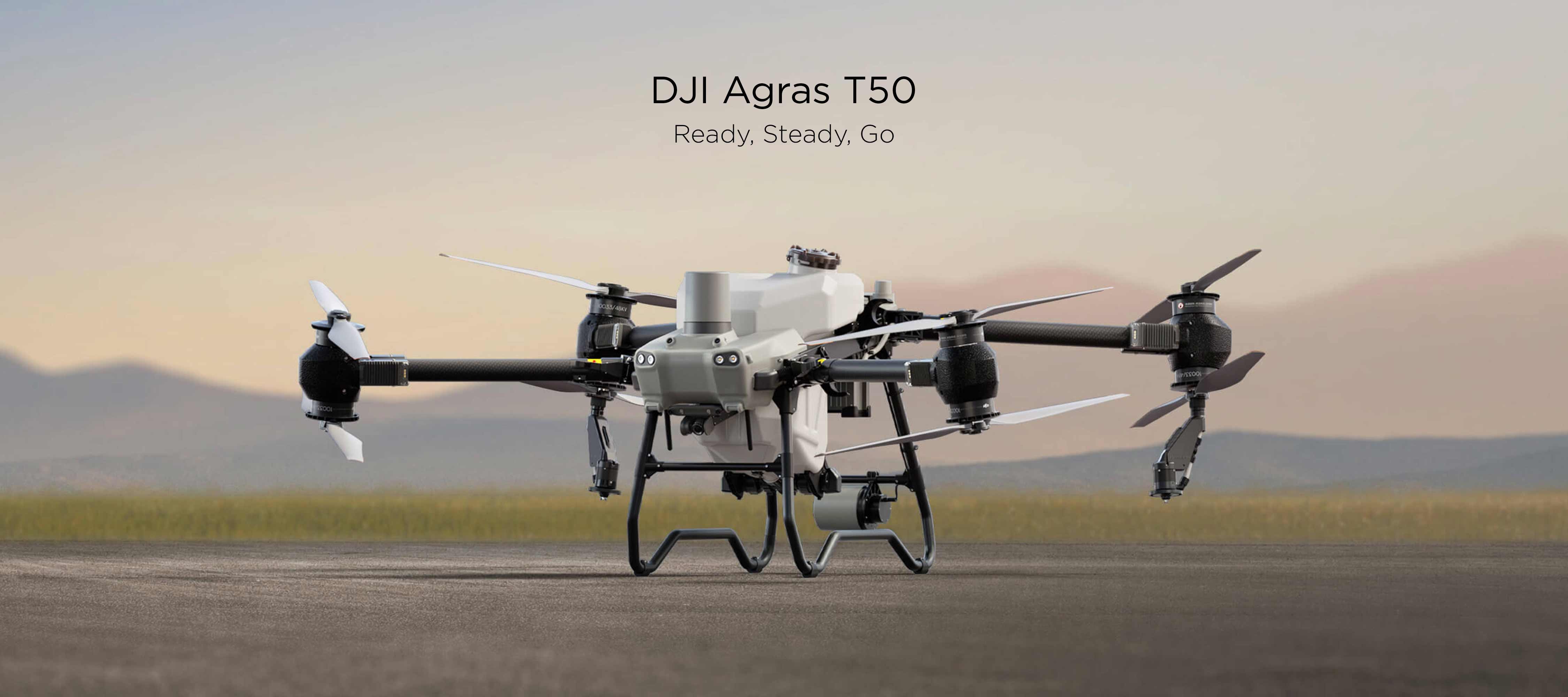 The DJI Agras T50 rests on top of sandy terrain with out-of-focus grass and mountains in the background.