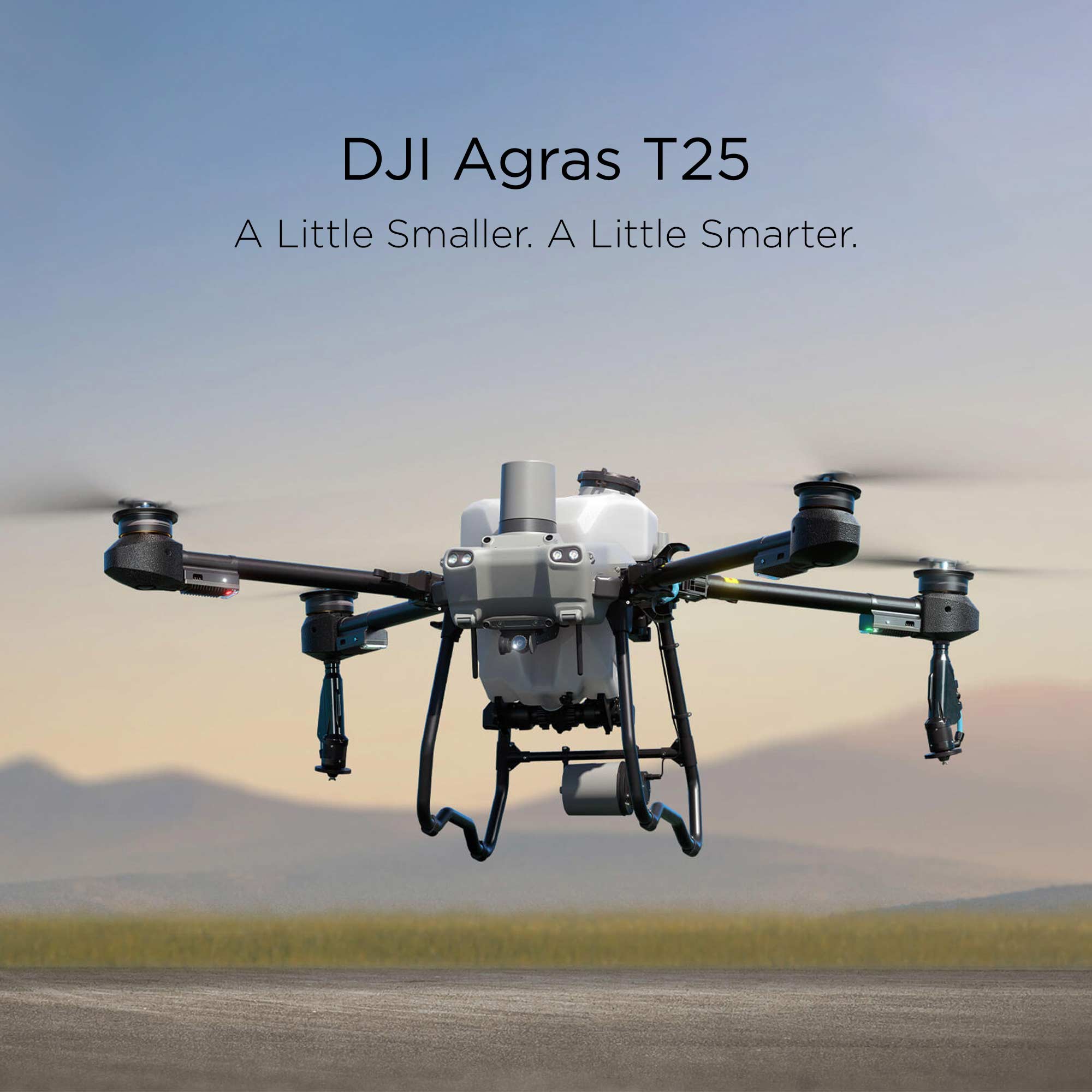 The DJI Agras T25 hovering over sandy terrain with out-of-focus grass and mountains in the background.