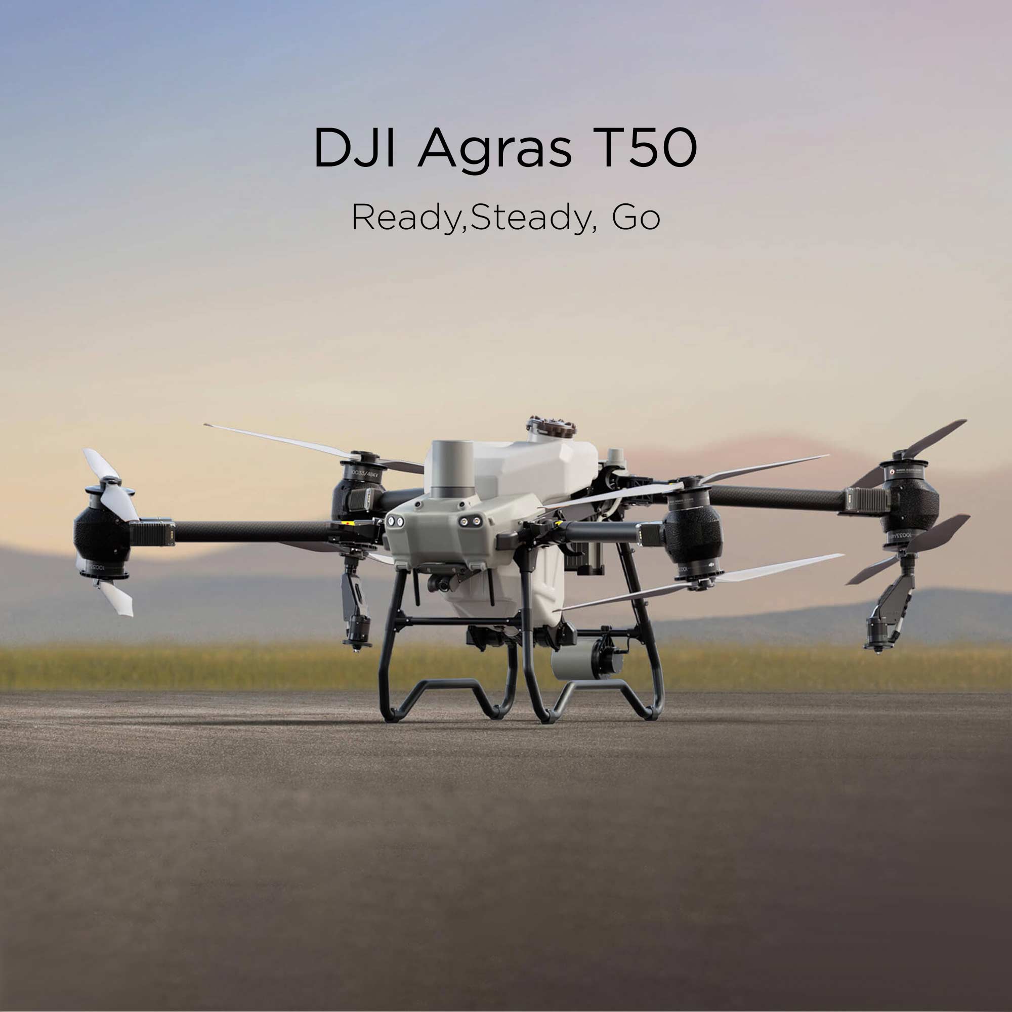 The DJI Agras T50 rests upon sandy terrain with out-of-focus grass and mountains in the background.